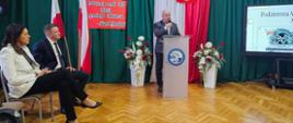 Mężczyzna w szarym garniturze przemawia do mikrofonu, stojąc za przezroczystą mównicą z logo szkoły (ZSA). W tle znajduje się dekoracja w barwach narodowych: zielone zasłony, polska flaga, kontur mapy Polski oraz czerwona tablica z cytatem Józefa Piłsudskiego: „Podstawą armii jest DUSZA prostego żołnierza...”. Po lewej stronie siedzą zaproszeni goście, w tym kobieta w białym żakiecie i mężczyzna w garniturze. Po prawej widać ekran z prezentacją o tytule „Podziemna Armia”.