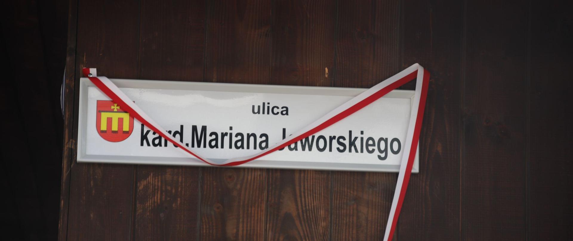 Na zdjęciu: tablica z napisem ulica kard. Mariana Jaworskiego, na drewnianych deskach domu opleciona biało-czerwoną szarfą.