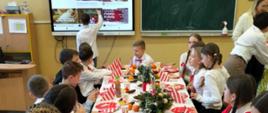 Przy stole nakrytym białym obrusem i czerwonymi serwetkami oraz zielonymi stroikami siedzą uczniowie i uczennice, spożywają przygotowany dla nich poczęstunek wigilijny