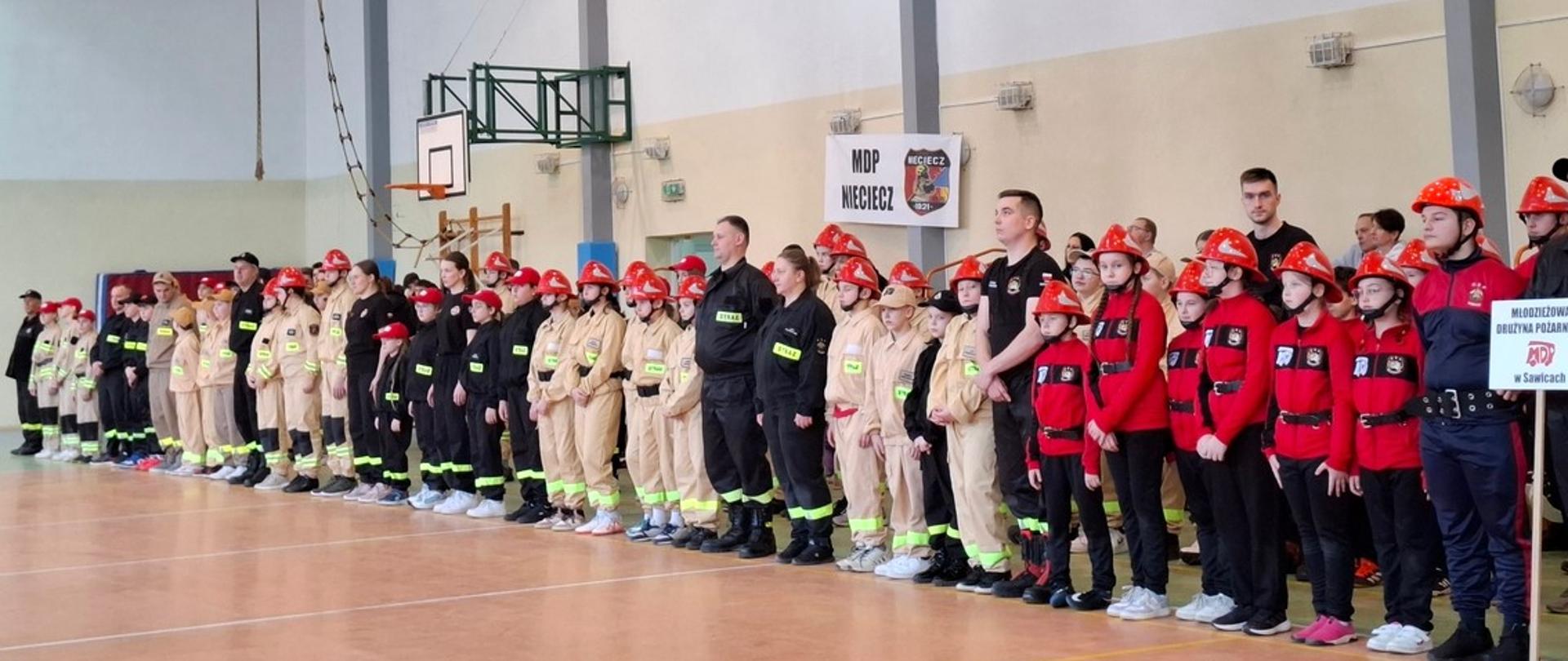 Fotografia wykonana podczas I Powiatowych Halowych Zawodów Sportowo – Pożarniczych MDP na hali sportowej w Zespole Szkół Im. H. Mniszek w Sabniach. Drużyny Młodzieżowych Drużyn Pożarniczych stoją w rzędzie w jednakowych dresach pożarniczych - beżowych, czarnych i czerwonych, na głowie maja czerwone hełmy strażackie.