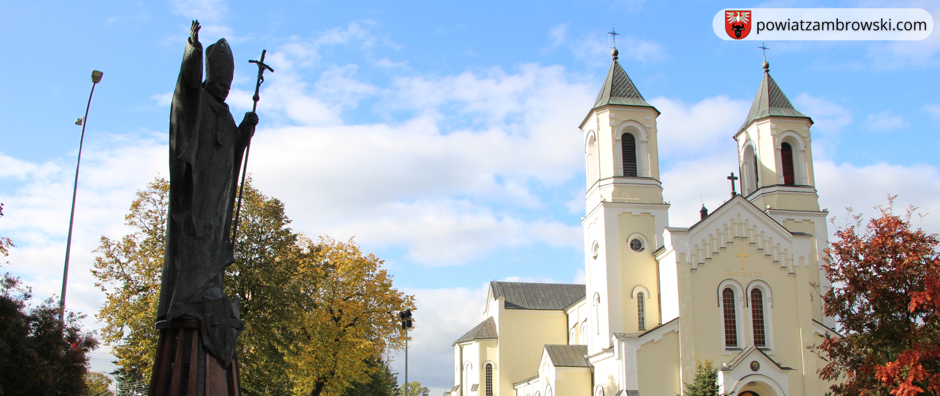 z lewej strony na zdjęciu znajduje się pomnik Jana Pawła II, w tle widoczny jest kościół pw. Trójcy Przenajświętszej w Zambrowie