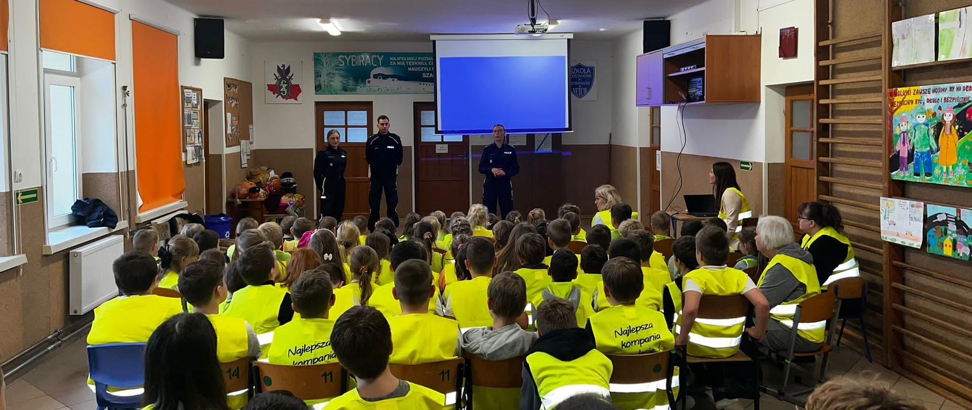 Na środku korytarza stoi troje funkcjonariuszy policji - dwie kobiety i mężczyzna. Ubrani są w mundury policyjne. Przed nimi na ławeczkach i krzesłach siedzą wszyscy uczniowie ubrani w kamizelki odblaskowe.