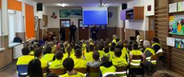 Na środku korytarza stoi troje funkcjonariuszy policji - dwie kobiety i mężczyzna. Ubrani są w mundury policyjne. Przed nimi na ławeczkach i krzesłach siedzą wszyscy uczniowie ubrani w kamizelki odblaskowe.