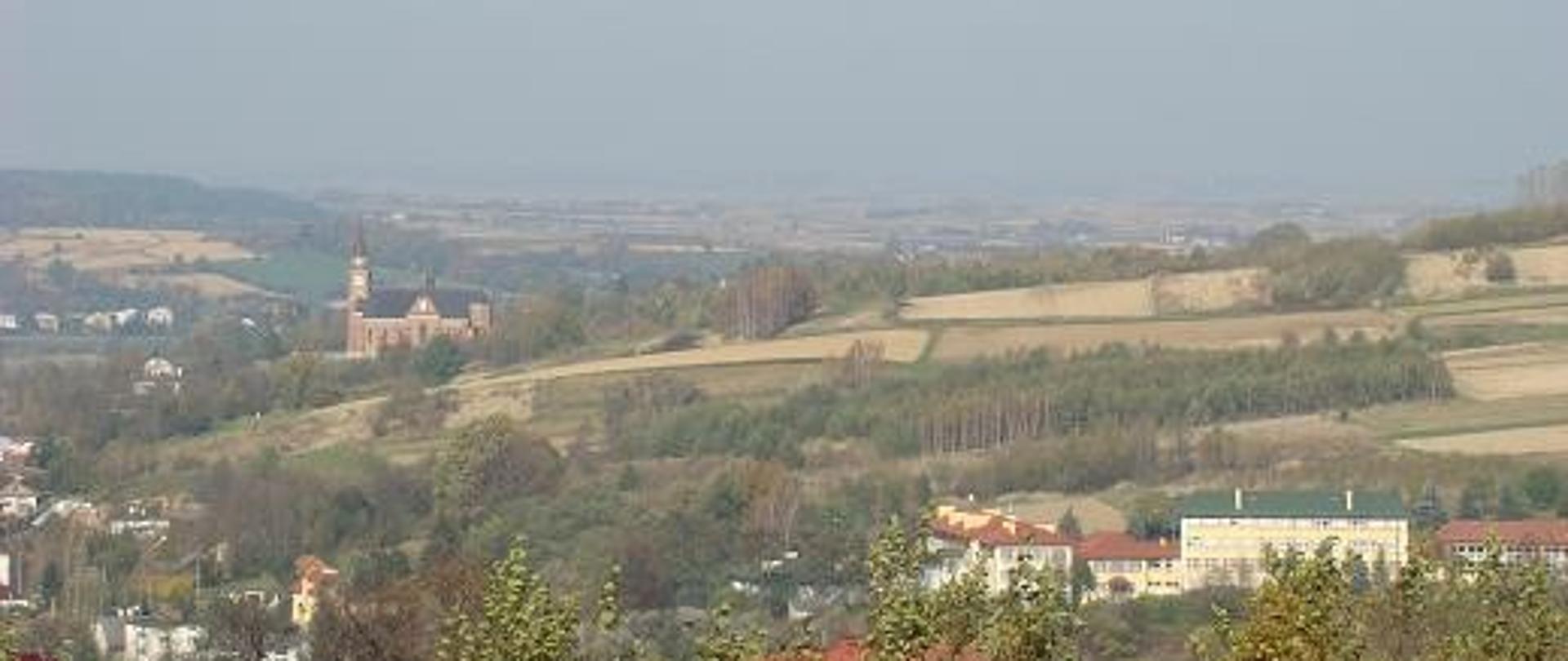 zdjęcie widokowe widoczna część miejscowości Lubenia w tle zbocze z kościołem w tle widoczna szkoła w Lubeni zabudowania pola uprawne i zadrzewienia widoczny horyzont z przyległymi gminami