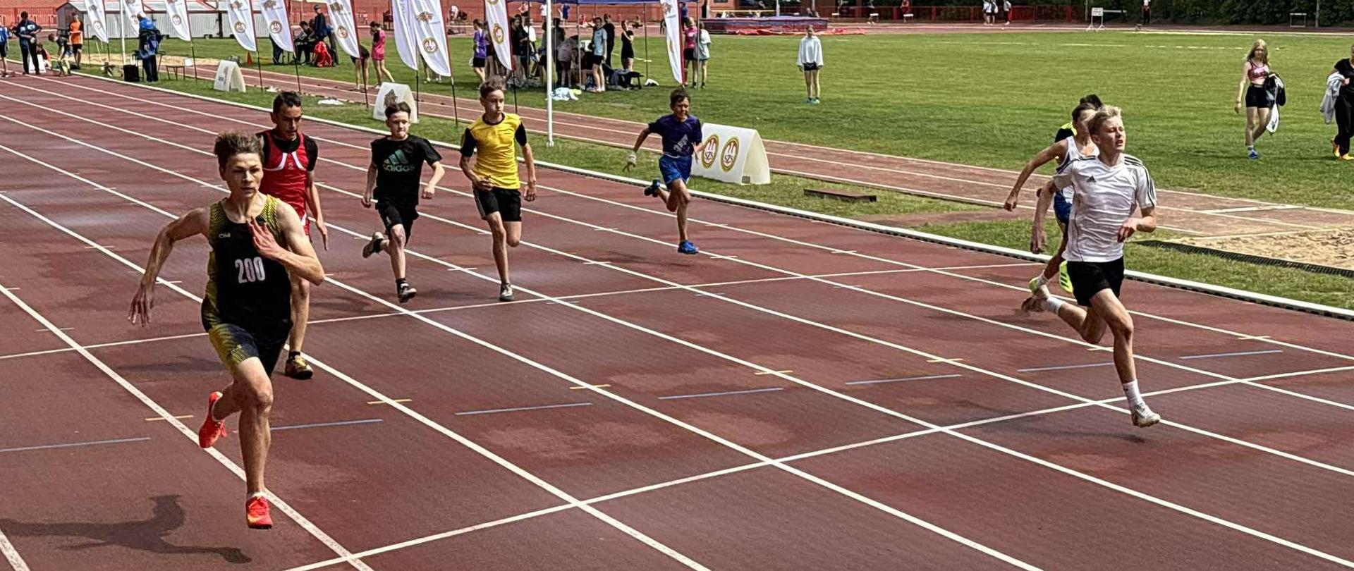Ośmiu chłopców ubranych w stroje sportowe biegnie po bieżni stadionu lekkokoatletycznego.