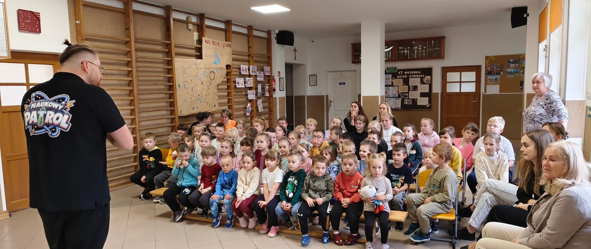 Mężczyzna w czarnym t-shircie z kolorowym logo „Naukowy Patrol” stoi tyłem do obiektywu, przemawiając do dużej grupy dzieci siedzących na ławkach i krzesłach w szkolnym korytarzu. 