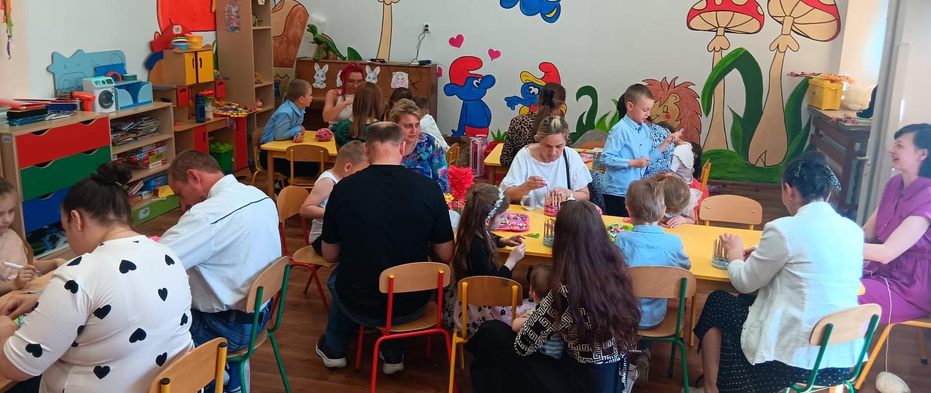 Rodzice siedzą przy stolikach w sali przedszkolnej i wykonują z włóczki ozdobne wazoniki, pomagają im dzieci