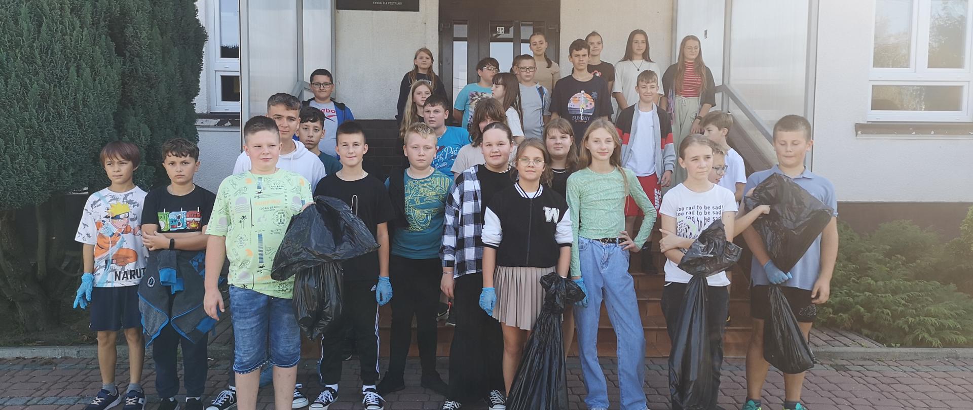 Uczniowie stoją na schodach przed budynkiem szkoły, kilkoro uczniów trzyma w rękach czarne worki