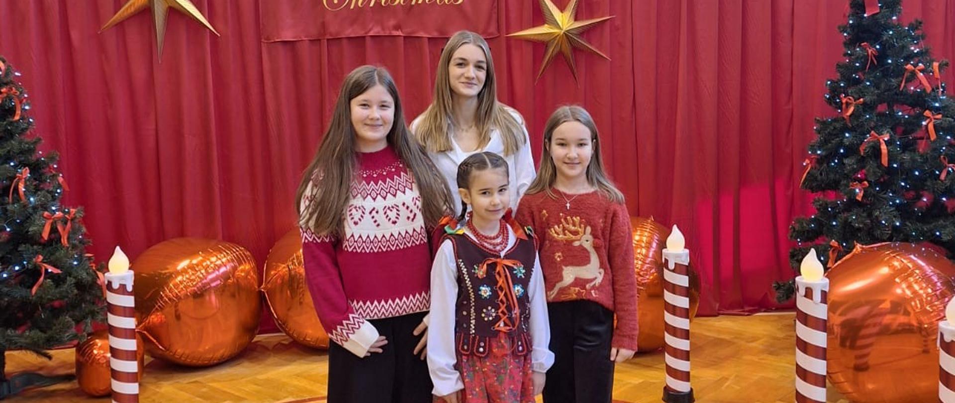 Trzy dziewczynki w Świątecznych sweterkach oraz jedna w gorsecie góralskim stoją po środku sali gimnastycznej pięknie udekorowanej świątecznie ( są dwie choinki, ogromne złote gwiazdy wiszą na bordowym tle, wielkie złote kule leżą na ziemi)
