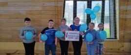 Uczniowie klasy trzeciej ubrani na niebiesko, trzymając niebieskie balony stoją na sali gimnastycznej solidaryzując się tym gestem z osobami będącymi w spektrum autyzmu