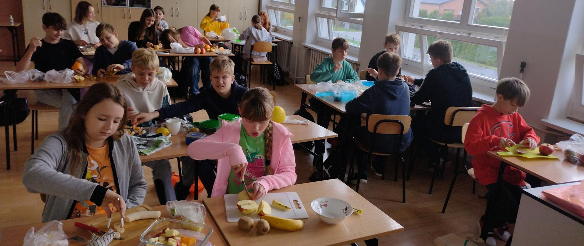 Przy stolikach siedzą uczniowie. Każdy z nich wykonuje sałatkę z przyniesionych przez siebie owoców.