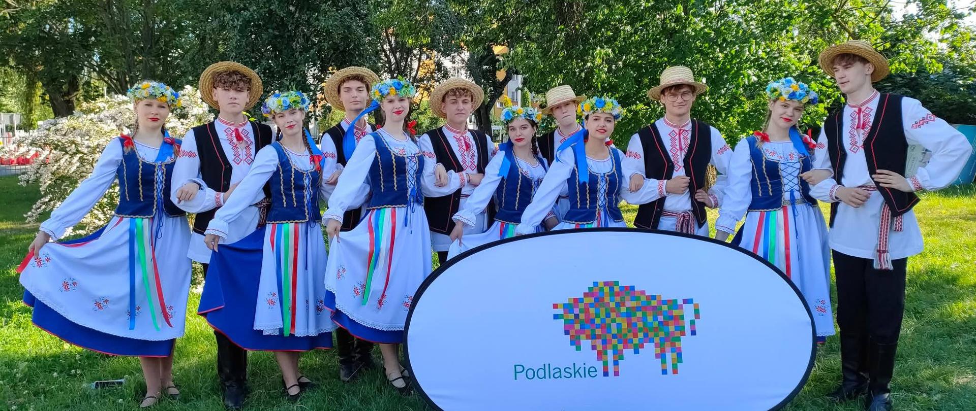 Młodzież prezentuje stroje ludowe, na tle baneru z logo Województwa Podlaskiego
