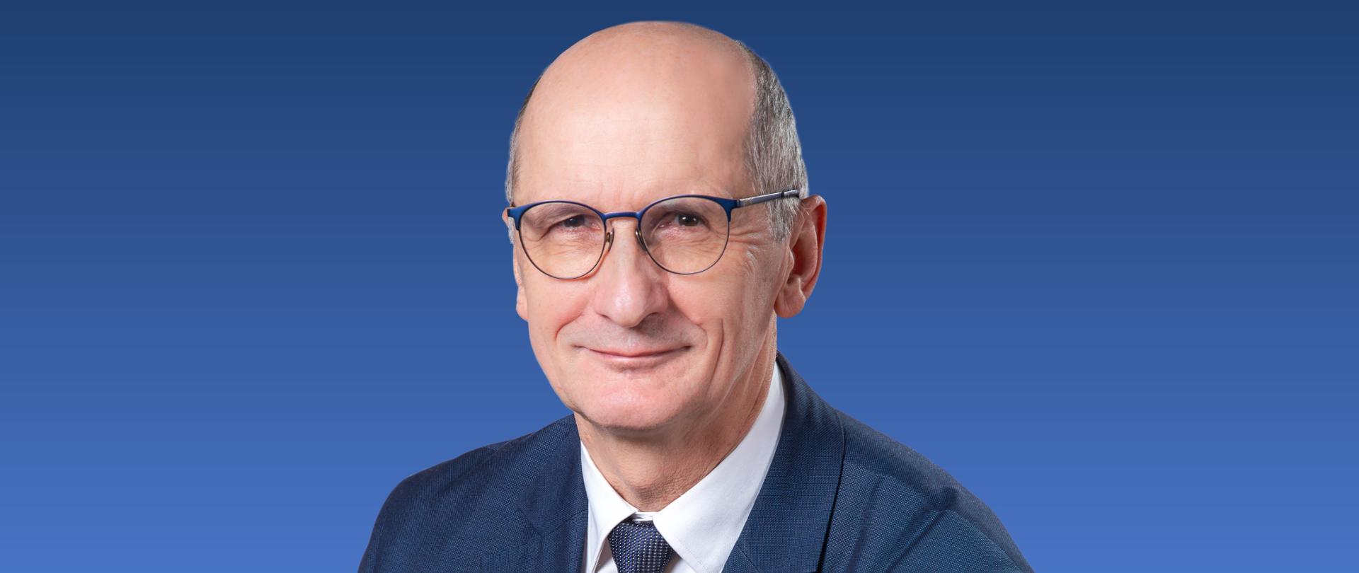 Zdjęcie przedstawia mężczyznę w okularach i w krawacie na niebieskim tle.