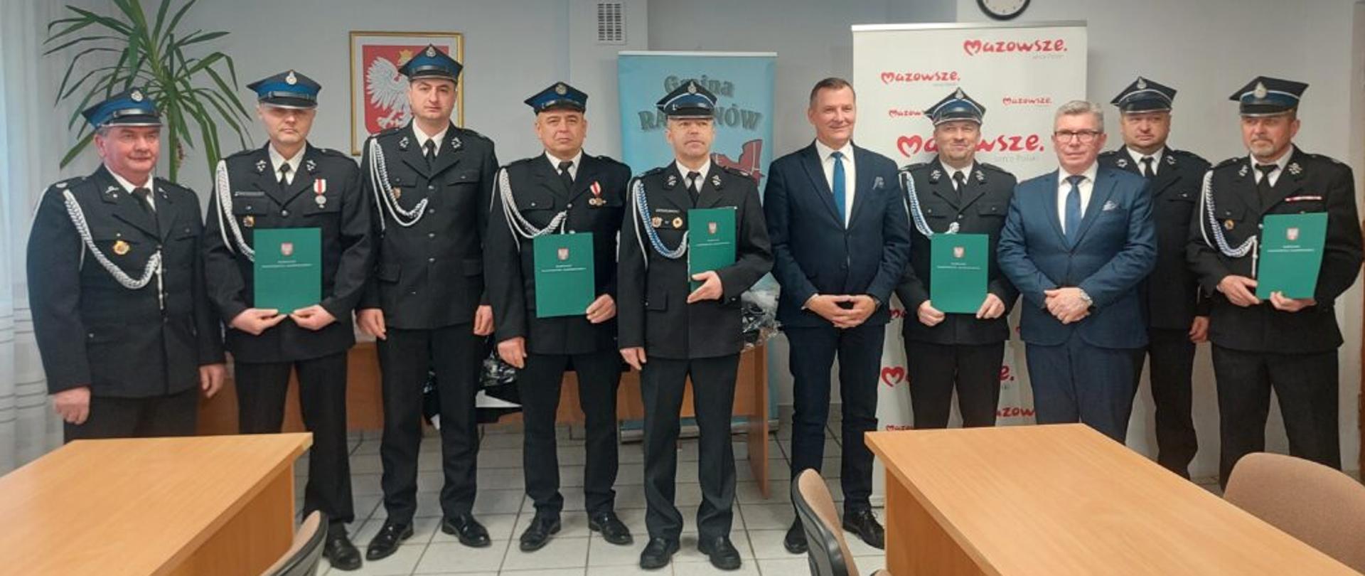 Grupa mężczyzn, niektórzy w formalnych mundurach i inni w garniturach, stoją w pomieszczeniu trzymając zielone teczki, pozując do zdjęcia