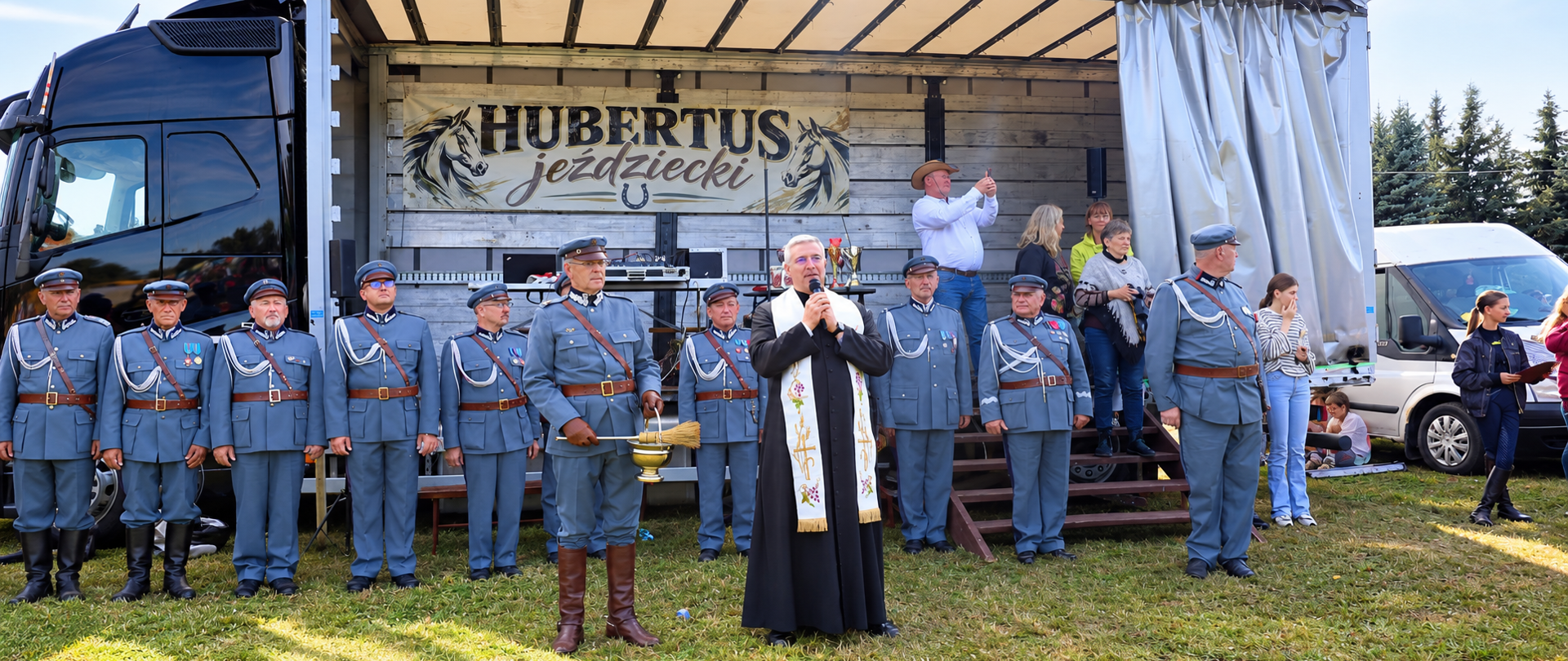 Uroczystość podczas Hubertusa Jeździeckiego. Na pierwszym planie ksiądz w stroju liturgicznym przemawia do zgromadzonych, a obok niego stoi mężczyzna w historycznym mundurze z kropidłem i naczyniem na wodę święconą. W tle widoczni są pozostali umundurowani uczestnicy oraz scena z banerem „Hubertus Jeździecki