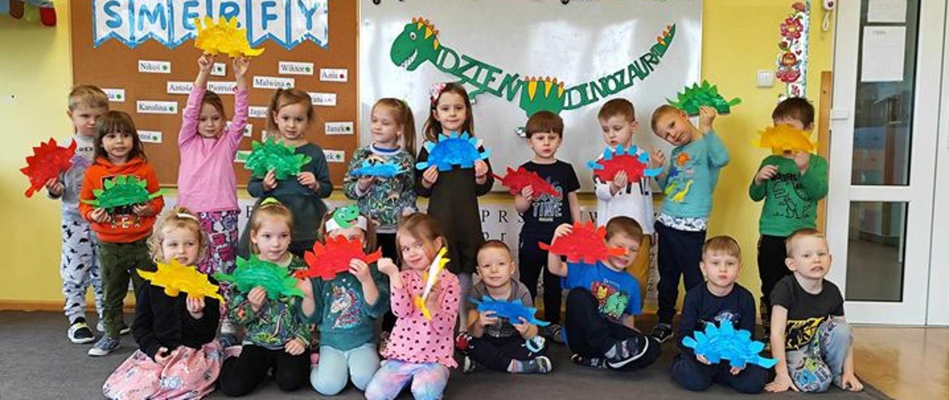 Grupa dzieci trzymająca samodzielnie wykonane papierowe stegozaury. W tle napis ‘Dzień Dinozaura’