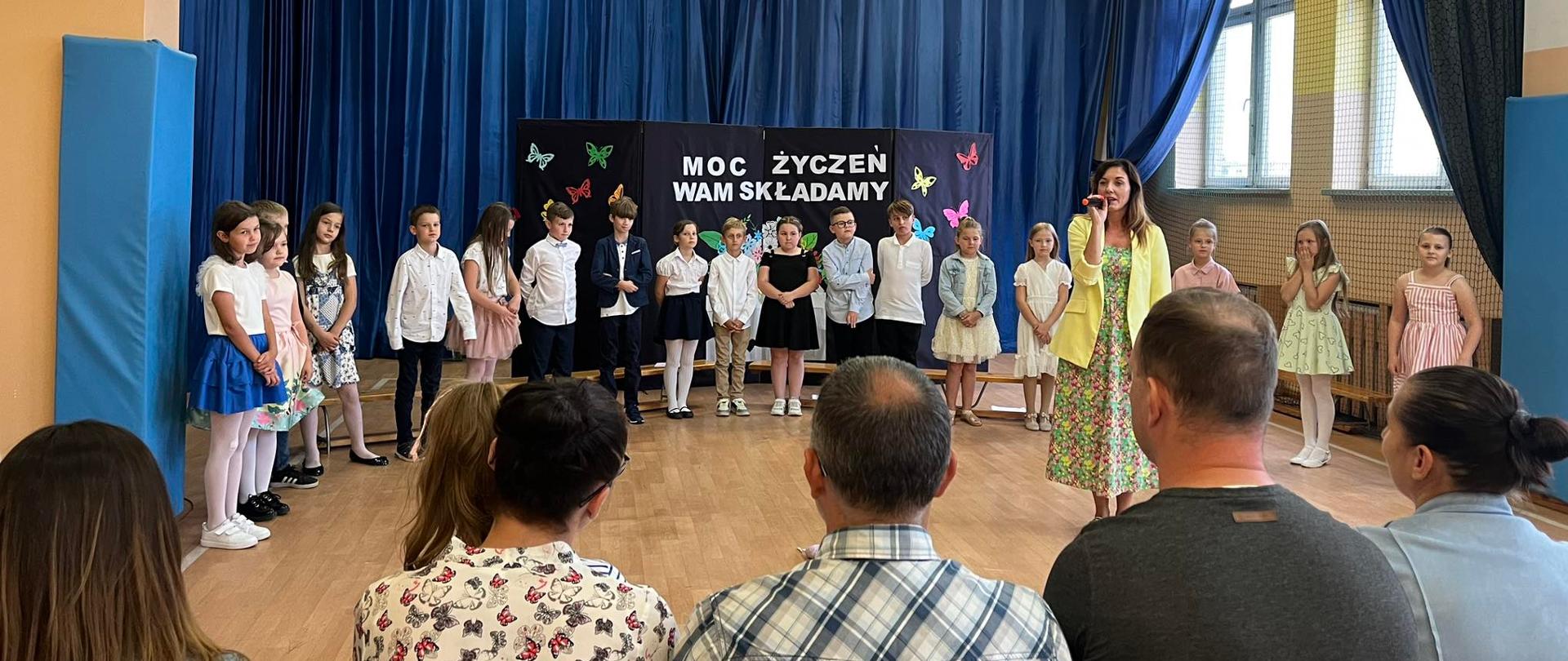 Dzieci ustawione obok siebie ubrane odświętnie.Za nimi na ścianie tekst Moc życzeń wam składamy. Przed nimi siedzą osoby dorosłe