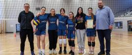 Fotografia na hali sportowej na tle siatki, sześć dziewcząt w ciemnoniebieskich koszulkach, jedna trzyma w ręku piłkę, druga dyplom, cztery pozostałe są między nimi, po lewej stronie sędzia zawodów, po prawej przewodniczący Rady Powiatu Sokołowskiego,