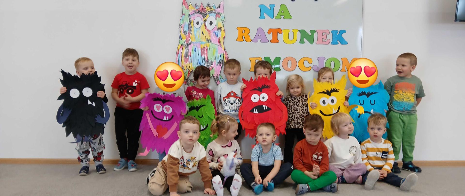 Grupa dzieci trzymająca kolorowe potwory na tle napisu „Na ratunek emocjom” i rysunku kolorowego potwora.