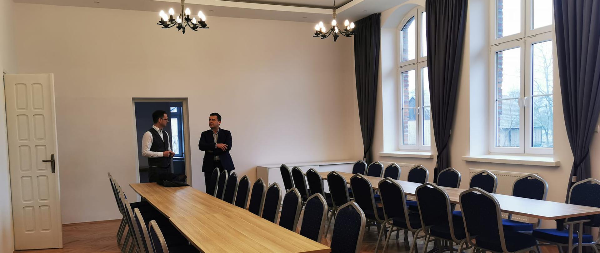 zdjęcie przedstawia odnowioną salę główną w budynku Domu Ludowego w Demblinie. W sali znajdują się dwa rzędy ze stołami oraz ustawionymi obok nich krzesłami. na drugim planie obok wejścia stoją Wójt Gminy Wietrzychowice oraz Wiceprzewodniczący Sejmiku Województwa Małopolskiego