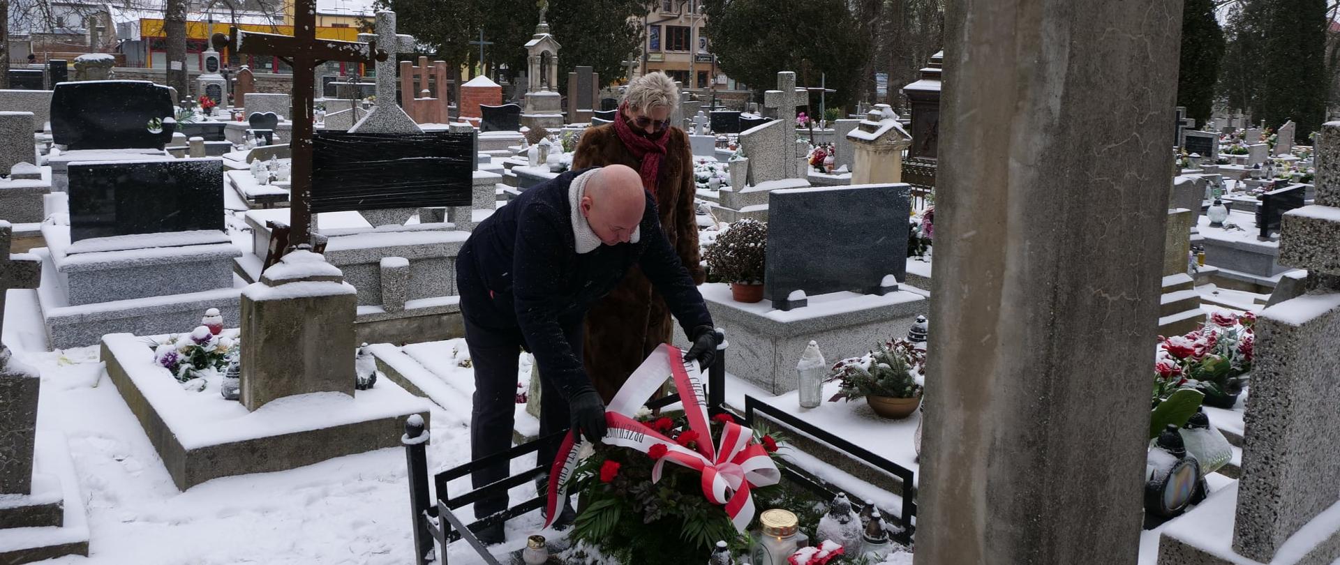 Zdjęcie przedstawia dwie osoby stojące na cmentarzu przed grobem w zimie. Jedna z nich kładzie na grobie wieniec.