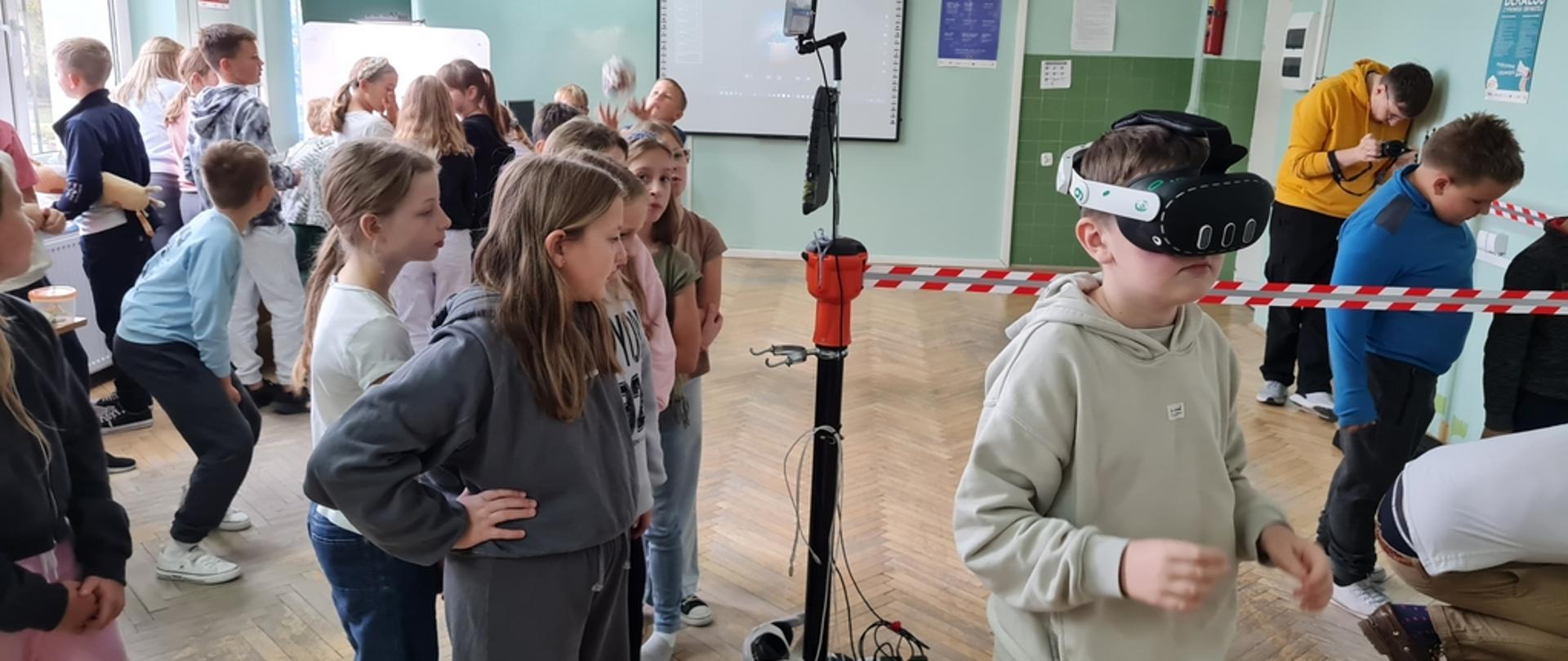 W szkolnej sali stojący chłopiec z założonymi na oczach okularami VR. Wokół niego przyglądające się dzieci.