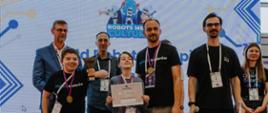 Zdjęcie grupowe na scenie podczas uroczystości wręczenia nagród zawodów robotycznych WRO (World Robot Olympiad). W tle ekran z logo „Robots Meet Culture". Na pierwszym planie dwoje dzieci z medalami na szyjach – chłopiec w czarnej koszulce z napisem „planeta koderów" trzyma puchar, drugi chłopiec w szarej bluzie trzyma dyplom z napisem „Super Wyszukiwarka". Za nimi stoją czterej dorośli mężczyźni oraz kobieta, część z nich ma na sobie koszulki z logo „planeta koderów" i „Open Source is Art". Wszyscy mają identyfikatory zawodów na smyczach.