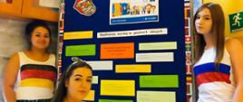 Europejski Dzień Języków. Na zdjęciu trzy nastolatki w koszulkach z flagami państw przy okolicznościowej tablicy na korytarzu szkolnym