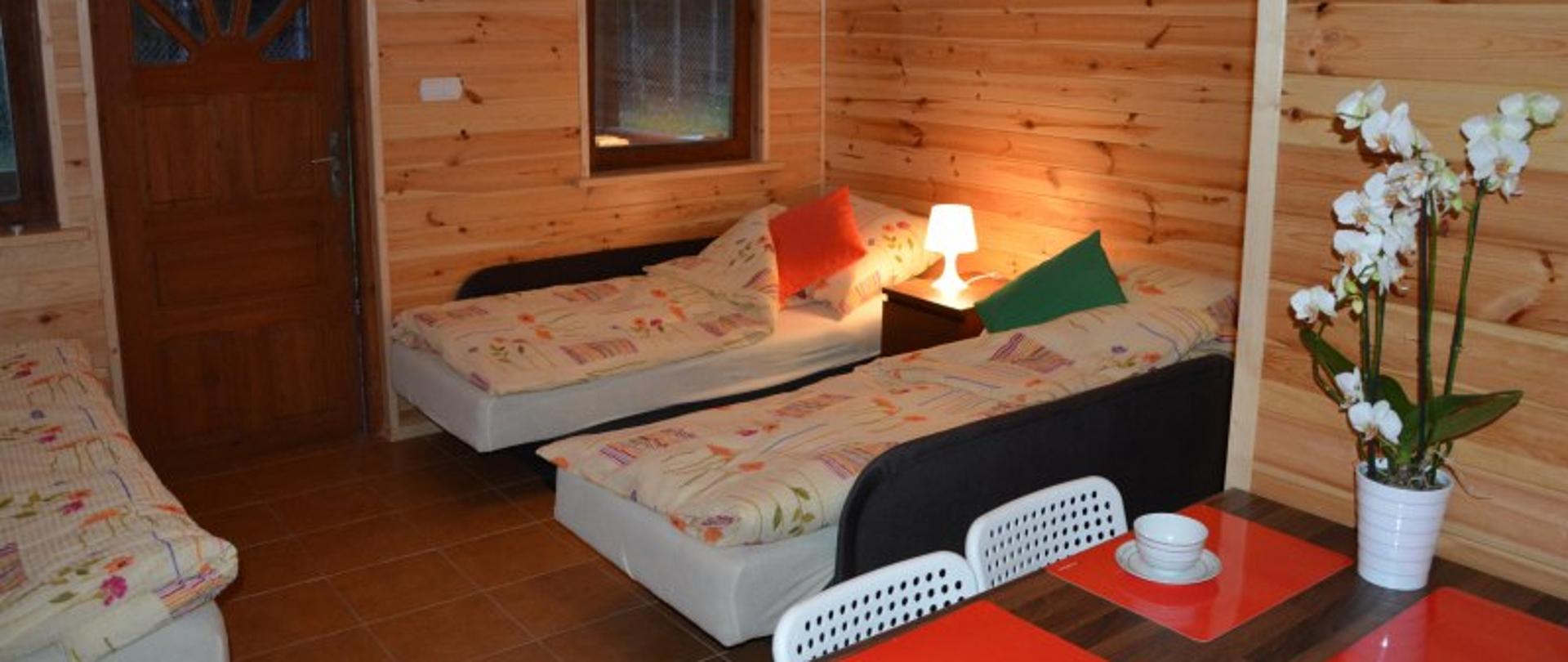 Zdjęcie wnętrza domku do wynajęcia. Ściany wyłożone drewnem, 3 pojedyncze łóżka, stół na którym stoi storczyk. Na podłodze płytki.