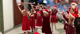 Na korytarzu w Szkole Filialnej ubrane na czerwono dziewczynki, w czerwonych czapkach, tańczą układ taneczny, podnoszą do góry ręce trzymające biało-czerwone laski, za nimi granatowa dekoracja przedstawiająca sanie z Mikołajem ciągnięte przez cztery renifery