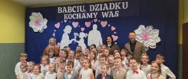 Wszystkie dzieci występujące na akademii stoją do zdjęcia razem z panią dyrektor, panią wójt i radnym gminy, w tle na granatowej zasłonie przypięte wycięte z białego papieru postaci babci i dziadka z wnuczką i wnukiem w ramce utworzonej z różowych serduszek, nad nimi napis "Babciu, dziadku, kochamy Was"