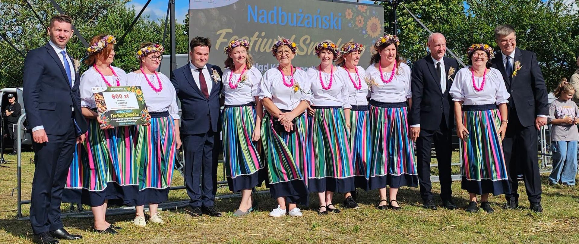 Grupa wykonawców w kolorowych spódnicach trzymająca certyfikat, pozująca z kilkoma mężczyznami w garniturach podczas wydarzenia plenerowego.
