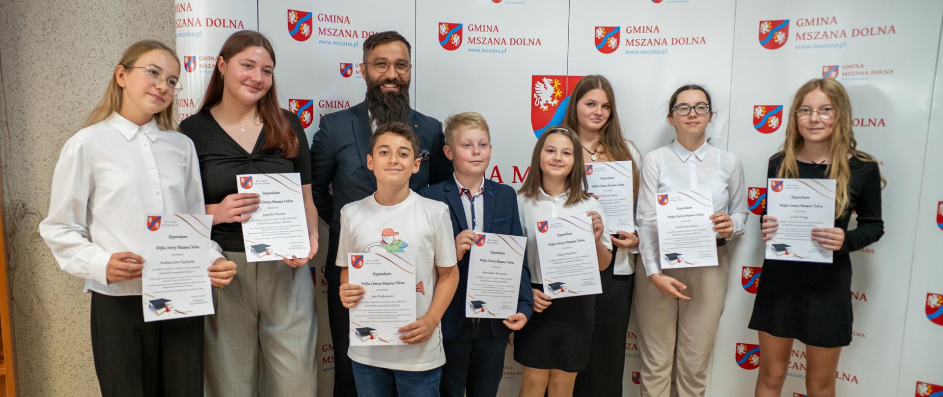 Na ściance zdjęciowej ustawiona grupa dzieci odświętnie ubranych wraz z Wójtem gminy. W ręku trzymają dyplomy.