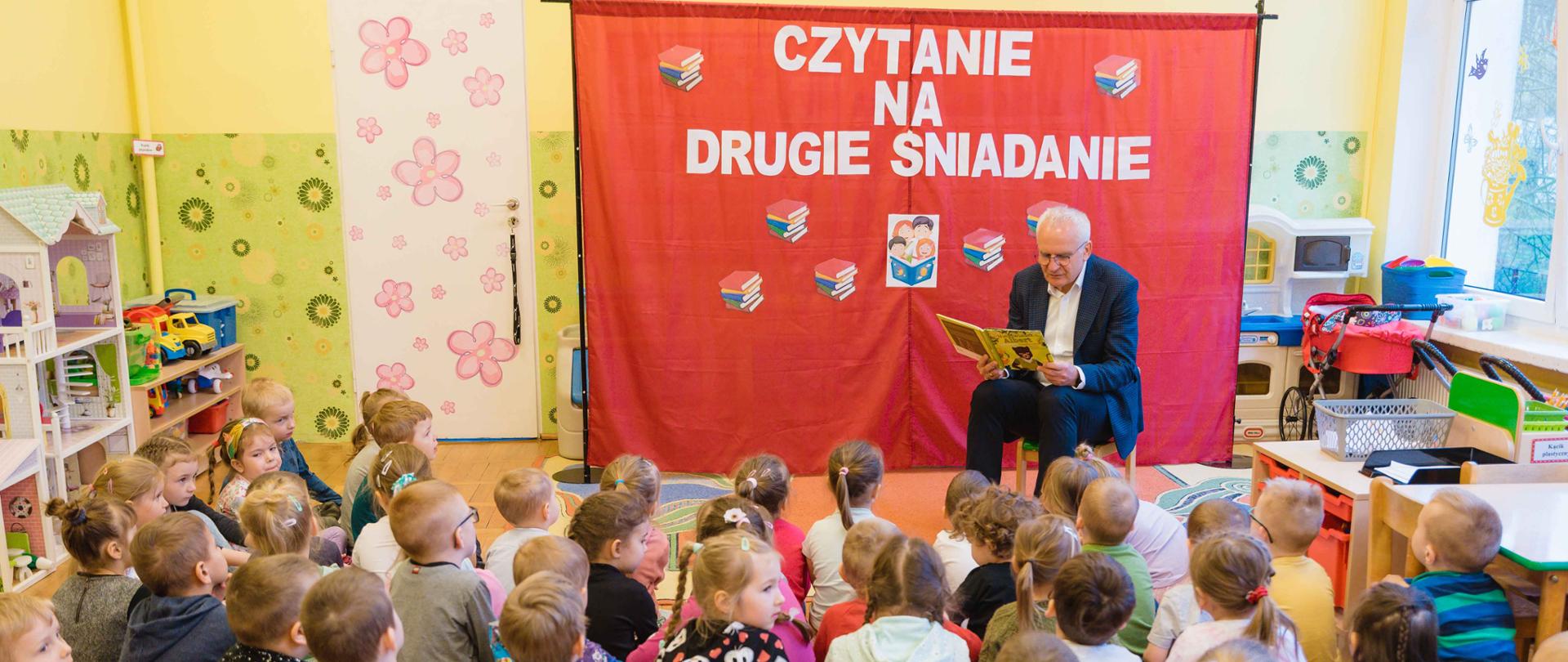 mężczyzna w garniturze czyta przedszkolakom książkę, dzieci siedzą na podłodze