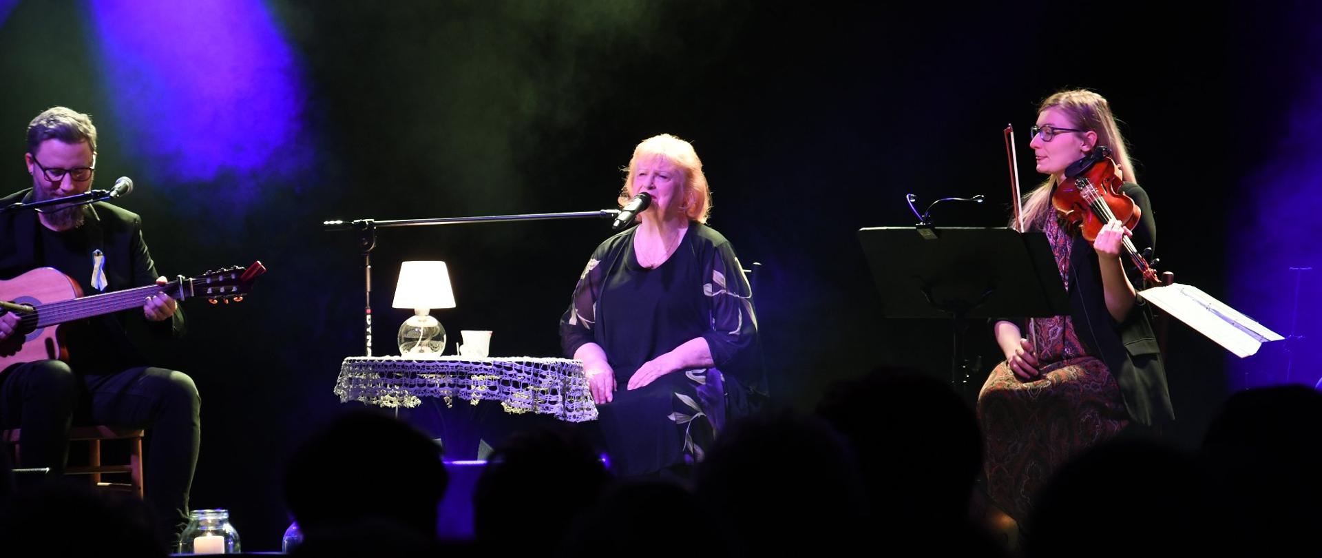 Trzy osoby siedzące na scenie, Stanisława Celińska oraz dwoje muzyków, kobieta grająca na skrzypcach i mężczyzna grający na gitarze. Przy Stanisławie stolik z lampką. Zdjęcie zrobione w trakcie koncertu.