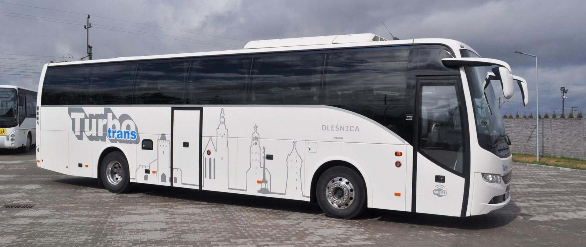 Biały autobus z logo przewoźnika stoi na parkingu, placu, wybrukowanym kostką