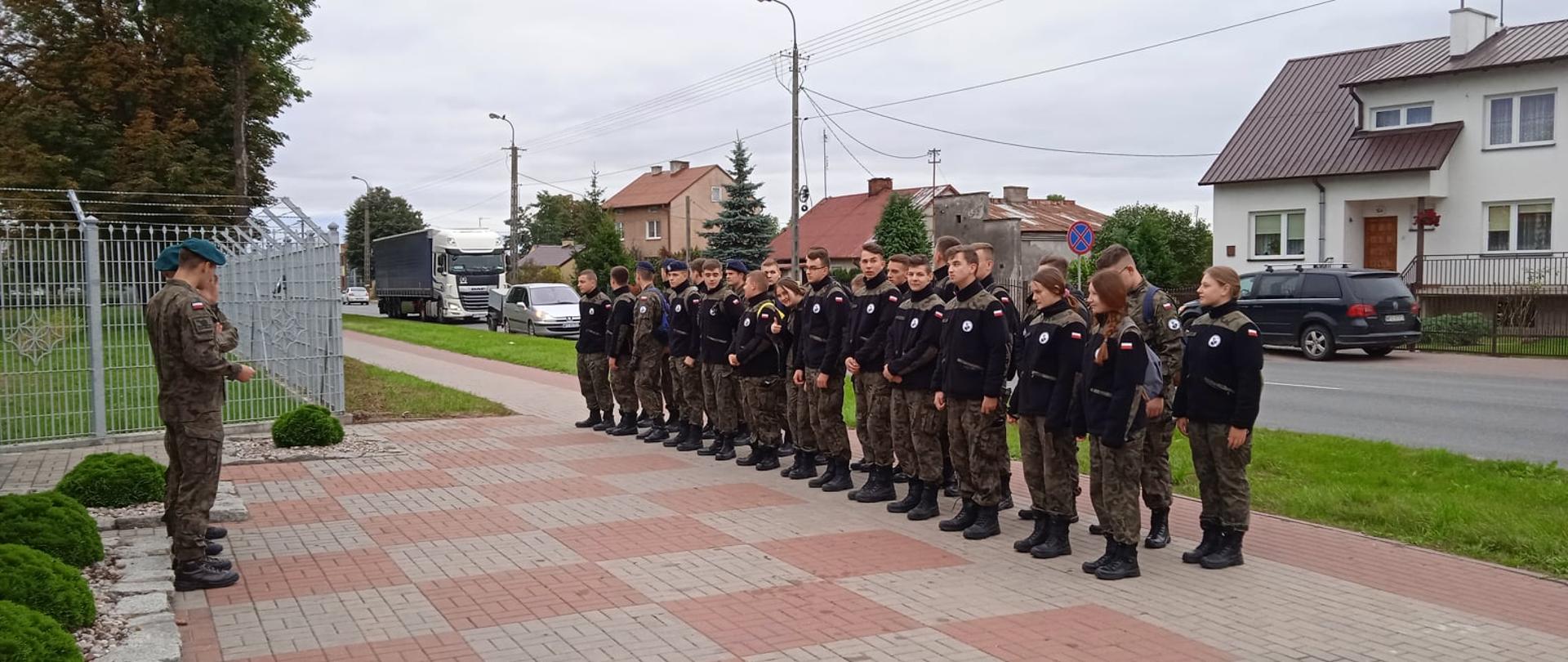 Szkolenie klas mundurowych Zespołu Szkół Powiatowych w Przasnyszu. Fotografia przedstawia uczniów klas mundurowych Zespołu Szkół Powiatowych w Przasnyszu (po prawej) stojących w szeregu przed dwoma żołnierzami jednostki (po lewej). Zdjęcie wykonano przed głównym wejściem na teren Jednostki w Przasnyszu.