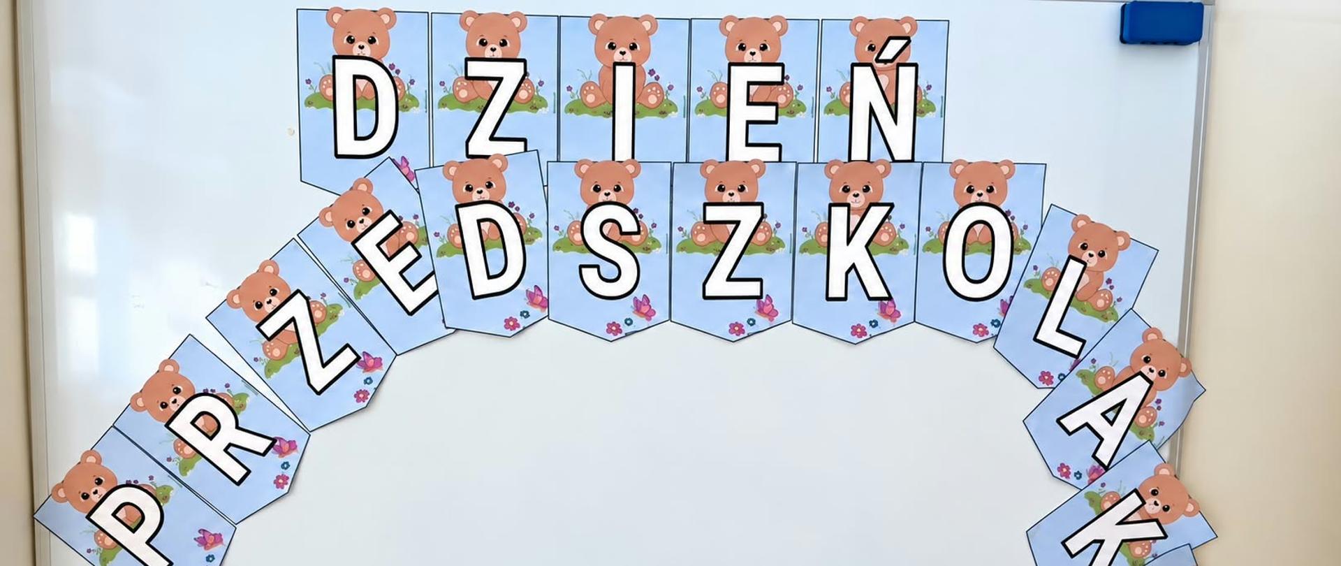 Biała tablica, na niej przyklejony napis Dzień Przedszkolaka. Każda litera jest na oddzielnej niebieskiej kartce, nad literami znajdują się misie.