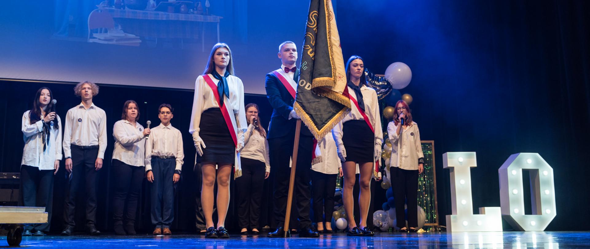 Na fotografii moment odśpiewania hymnu szkoły. Na pierwszym planie poczet sztandarowy składający się z dwóch uczennic oraz ucznia. W tle grupa uczniów ustawiona w szeregu, którzy śpiewają hymn. 