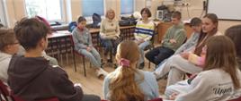 Uczniowie siedzą na krzesłach ustawionych w kole w swojej klasie, między nimi siedzi pani psycholog prowadząca zajęcia i nauczycielka języka polskiego