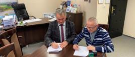 Burmistrz Margonina Janusz Piechocki i proboszcz parafii w Margoninie Rafał Wnuk podpisują dokument siedząc przy brązowym stole w gabinecie burmistrza