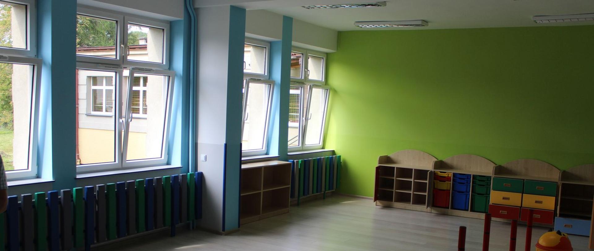 Na zdjęciu wnętrze sali przedszkola w Barwałdzie Górnym. Widok na okna i zieloną ścianę i wielokolorowymi meblami. 