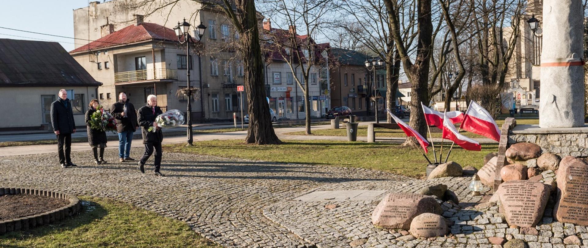 Wiceminister zdrowia, senator RP Waldemar Kraska podchodzi z wiązanka kwiatów do pomnika "Żołnierzy Niezłomnych" w kształcie krzyża z nałożoną żołnierską czapką ustawionego na kamiennej podstawie z licznymi kamieniami wokół, na których widnieją napisy upamiętniające daty, wydarzenia i osoby. Pomnik w Sokołowie Podlaskim został zbudowany w hołdzie żołnierzom Armii Krajowej i struktur poakowskich Obwodu Sokołów Podlaski, 6 Brygady Wileńskiej AK, kpt. 'Młota' oraz cywilnych mieszkańców powiatu sokołowskiego poległych w walce z komunistycznym zniewoleniem w latach 1944-1953. W tle powiewają biało czerwone flagi, widać skwer i elementy architektury miejskiej. Za senatorem stoją od lewej wicestarosta Ryszard Domański, starosta Elżbieta Sadowska, przewodniczący Rady Powiatu Sokołowskiego Jacek Odziemczyk.
