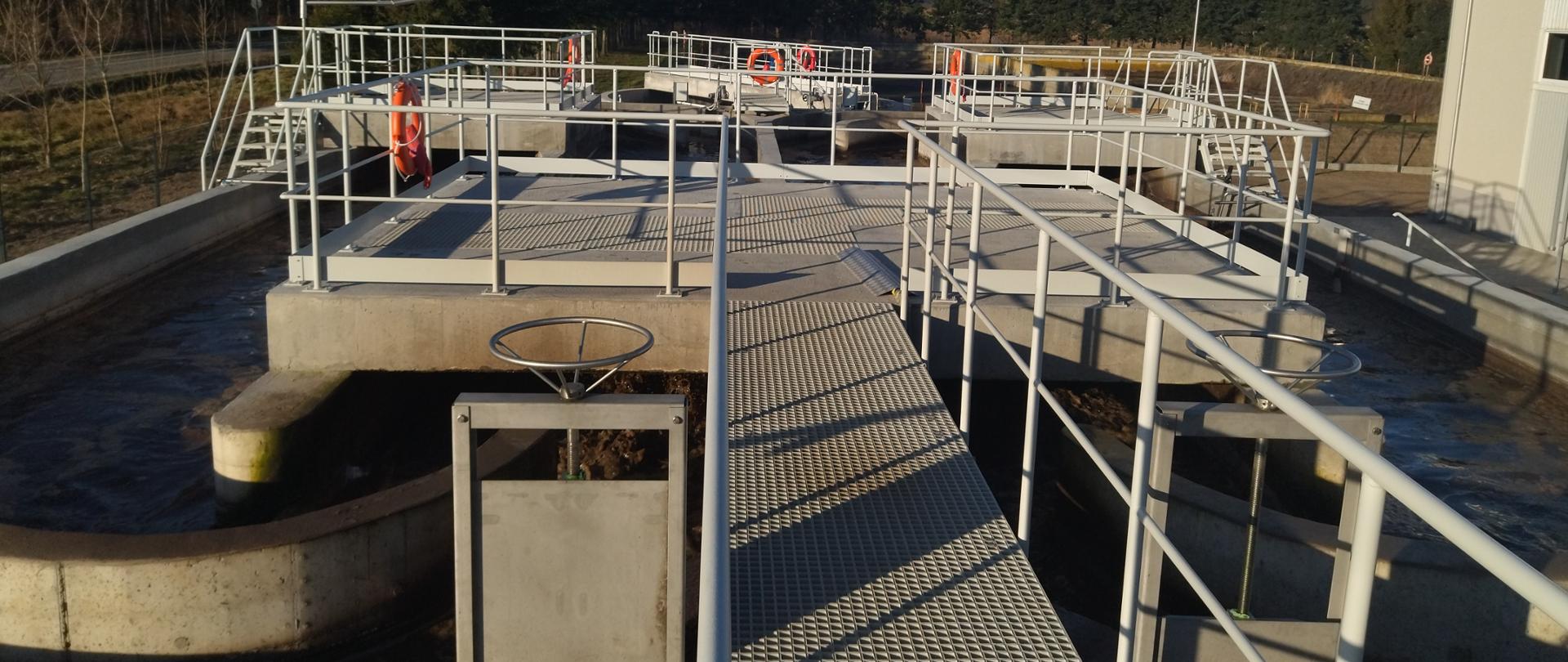 Na zdjęciu widoczna jest oczyszczalnia ścieków. W skład infrastruktury wchodzą betonowe zbiorniki oraz metalowe kładki z poręczami, do których przymocowane są pomarańczowe koła ratunkowe. W zbiornikach znajduje się woda, a na konstrukcji można dostrzec różne mechaniczne elementy, takie jak zawory i rury. Całość jest otoczona drzewami, a nad obiektem rozciąga się bezchmurne niebo. Obiekt znajduje się w obszarze o charakterze wiejskim lub podmiejskim i pełni istotną funkcję w gospodarce wodnej.