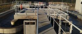 Na zdjęciu widoczna jest oczyszczalnia ścieków. W skład infrastruktury wchodzą betonowe zbiorniki oraz metalowe kładki z poręczami, do których przymocowane są pomarańczowe koła ratunkowe. W zbiornikach znajduje się woda, a na konstrukcji można dostrzec różne mechaniczne elementy, takie jak zawory i rury.
