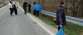 Uczniowie wędrują wzdłuż drogi i zbierają śmieci, jedna dziewczynka trzyma w ręku niebieski worek ze śmieciami