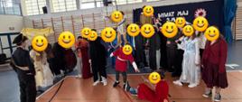 Na górze zdjęcia biała ściana z oknami, na których są białe rolety. Poniżej okien na białej ścianie rząd drabinek a na ich tle ciemny materiał z napisem z białych liter. Pośrodku zdjęcia sylwetki osób stojące w półkolu, ubrane w ciemne ubrania stojące. Pośrodku nich dwie osoby ubrane w stroje koloru czerwonego. Wszystkie osoby są na podłodze w kolorze jasnym brązowym.