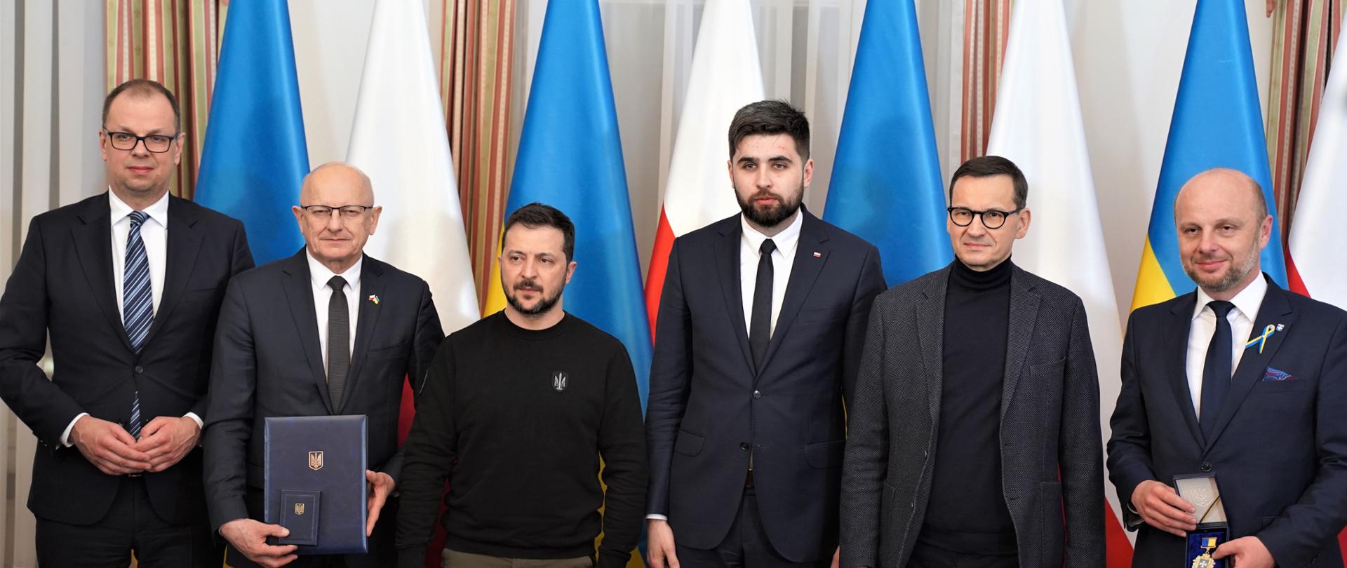 Zdjęcie przedstawia sześć osób stojących wewnątrz budynku na tle flag