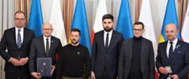 Zdjęcie przedstawia sześć osób stojących wewnątrz budynku na tle flag