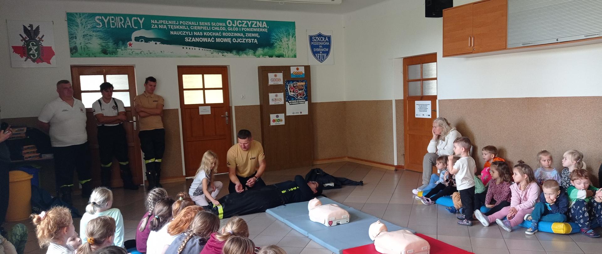 Na środku korytarza znajduje się mężczyzna ubrany w pomarańczową koszulkę z napisem: STRAŻ. Obok niego znajduje się dziewczynka. Oboje nachylają się nad kobietą leżącą na materacu.